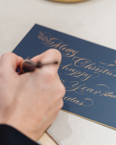 Eine Hand die eine Feder hält und mit goldener Tinte eine Karte beschriftet